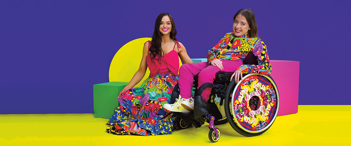 Photo of two people posing in colorful clothing and one in a colorful wheelchair